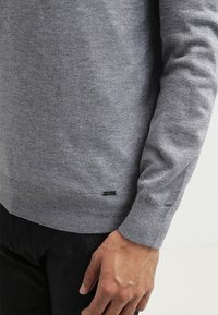 Gray knitted sweater with a ribbed hem and cuffs. Tag visible near bottom hem. Smooth texture. Part of a black pant is partially shown.