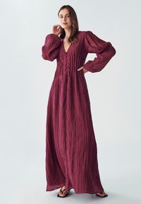 Dlouhá burgundská šaty s hlubokým výstřihem do V, knoflíkovým zapínáním vpředu a texturovanou tkaninou. Nabízí objemové rukávy a design až na podlahu.