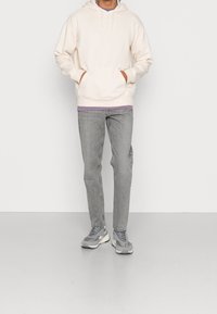 Světle béžová mikina s klokankou, pod ní levandulová košile, v kombinaci se šedými džínami a šedými teniskami. Hladká struktura látky.