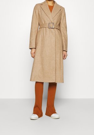 Kvinde iført en kamelfarvet uldfrakke med bælte over rustfarvede ribbukser og hvide sneakers, stående foran en ensfarvet hvid baggrund.