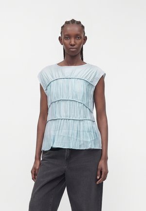 Femme aux cheveux tressés porte un haut plissé bleu clair sans manches et un pantalon noir ample, debout devant un fond blanc uni.