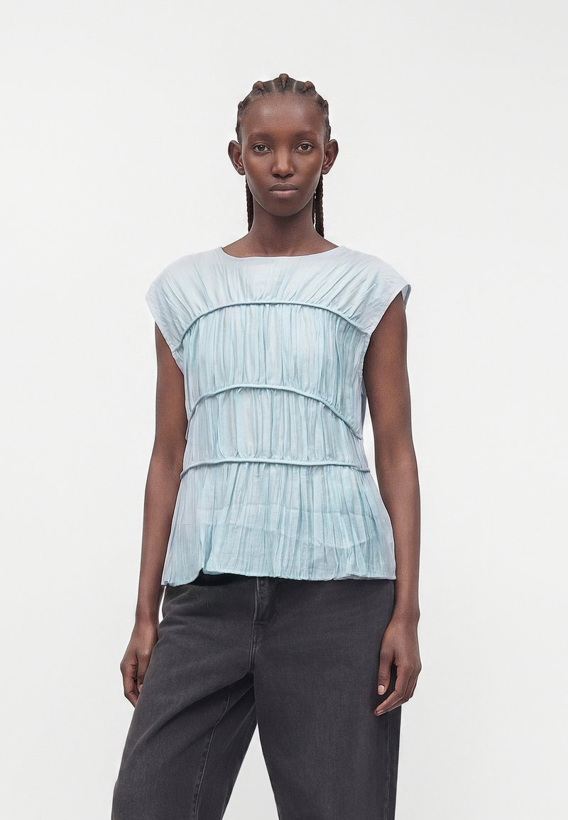 Femme aux cheveux tressés porte un haut plissé bleu clair sans manches et un pantalon noir ample, debout devant un fond blanc uni.