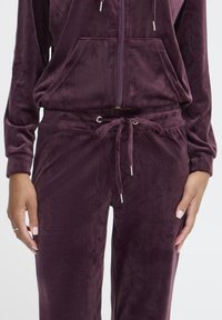 Aksamitny dres w głębokim bordowym kolorze z bluzą na suwak i przednimi kieszeniami. Spodnie mają ściągacz z troczkami i metalowe ozdoby. Miękka faktura na całej powierzchni.