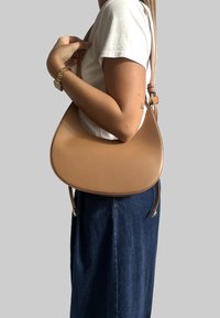 Torba na ramię w kolorze brązowym z zaokrąglonym kształtem, wyposażona w pojedynczy regulowany pasek i gładką teksturę. Noszona w zestawieniu z dżinsowym strojem.