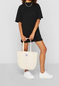 Beige canvas draagtas met een paarse label waarop een vlinderontwerp staat. De tas heeft lange hengsels en een rechthoekige vorm.