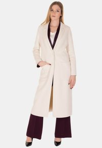 Cappotto lungo di colore crema con colletto a risvolto, due tasche frontali e foderato in seta. Abbinato a pantaloni bordeaux e tacchi neri.