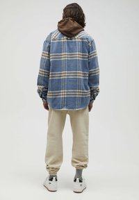 Geruite flanellen shirt in blauw, crèmekleurig en bruin, over een bruine hoodie gedragen. Gecombineerd met beige broek en witte sneakers met zwarte accenten.