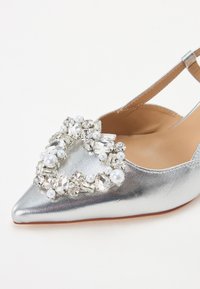 Zapato plano metálico plateado con punta afilada, decorado con cuentas transparentes y similares a perlas en forma de corazón en la parte frontal. Correa ajustable en el talón.