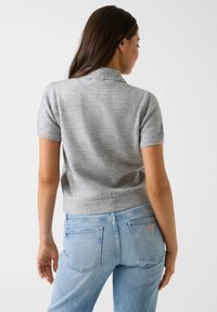 Pull gris à manches courtes avec col, en matière douce, associé à un jean bleu clair avec poches arrière et une étiquette logo.