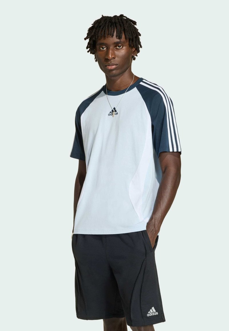 Męski model stoi z rękami w kieszeniach, ubrany w biało-granatową sportową koszulkę Adidas i czarne spodenki Adidas na jednolitym jasnym tle.