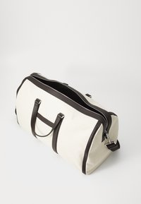 Borsa a sacca in tela crema con dettagli in pelle marrone scuro, dotata di doppi manici, chiusura con zip e tasca frontale.