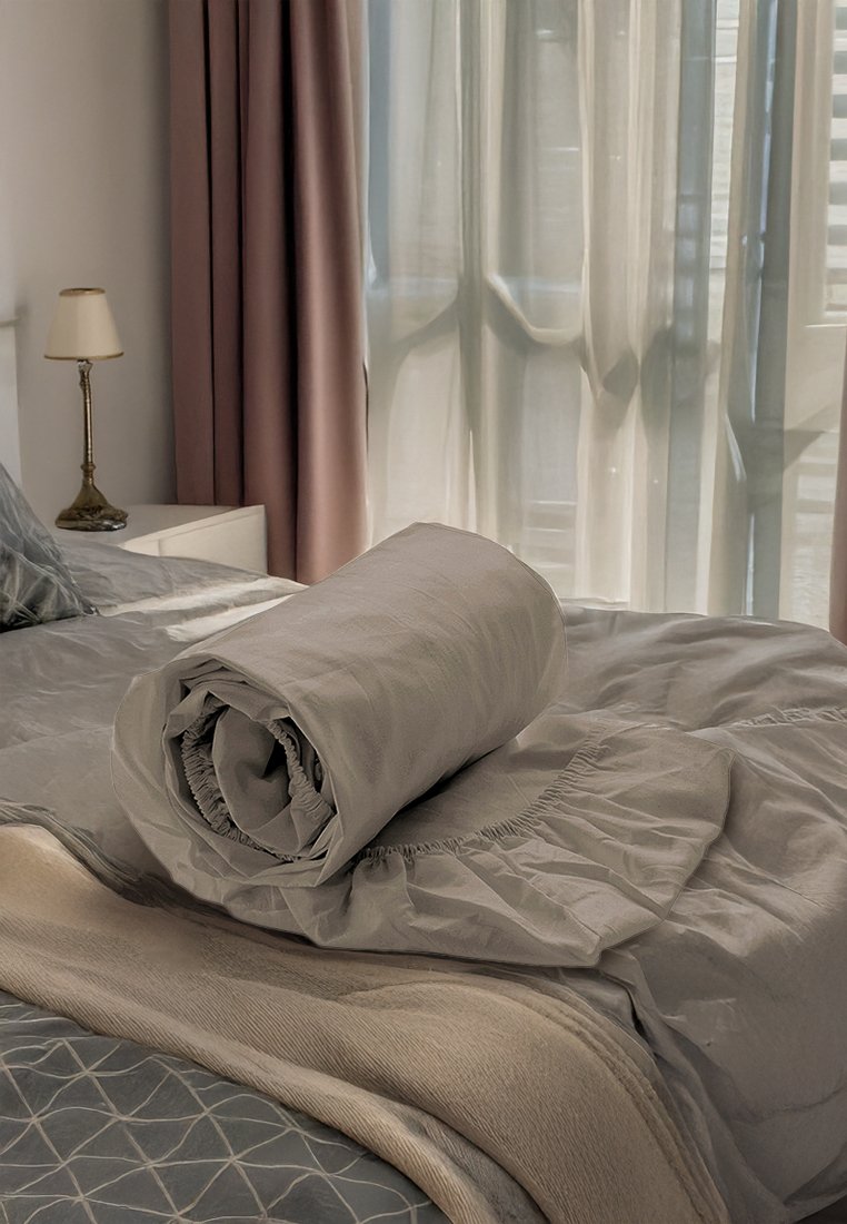 Grau gerollte Decke auf einem strukturierten beige- und graufarbenen Bettüberwurf. Weicher Stoff mit einem gerafften Rand, neben einer Lampe und transparenten Vorhängen.