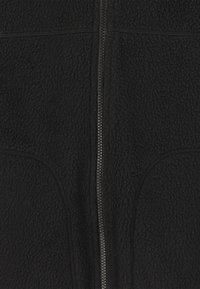Veste en polaire noire avec fermeture éclair sur le devant, deux poches latérales et tissu texturé. Le design met l'accent sur la chaleur et le confort.