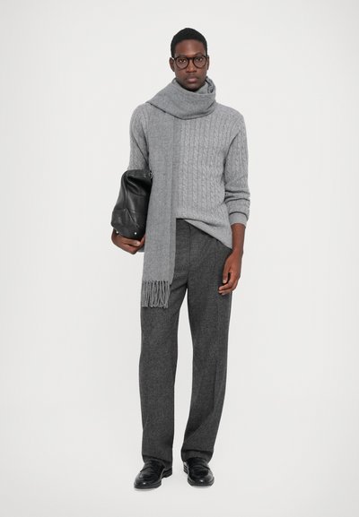 Grå kabelstrikket sweater med et matchende fransesjal, parret med grå bukser og sorte lædersko, holder en sort lædertaske.
