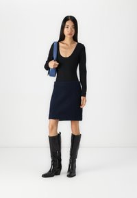 Čierny bodysuit s dlhým rukávom v kombinácii s námornícky modrou vlnou sukňou. Modelka nosí lesklé čierne čižmy po kolená a má so sebou modrú tašku na popruhu.