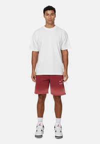 T-shirt bianco a maniche corte, pantaloni corti rossi con sfumatura e grafica, sneakers bianche e nere. Design semplice e vestibilità rilassata.