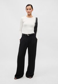 Fehér, hosszú ujjú felső, amit magas derekú fekete, bő szabású nadrággal párosítottak. Az öltözet fekete övet és egy vállon átdobott textúrált fekete kiegészítőt tartalmaz.