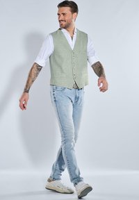 Mann in hellblauen Jeans, weißem, hochgekrempeltem Hemd, hellgrauem, geknöpftem Weste, weißen Sneakers, mit tätowierten Armen und lächelnd, vor einfarbigem Hintergrund.