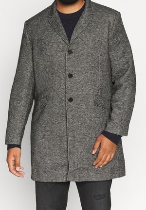 Mężczyzna ubrany w szary, teksturowany płaszcz do połowy długości z czarnymi guzikami, pod którym ma czarny sweter i ciemne spodnie, stoi na tle jednolitego tła.