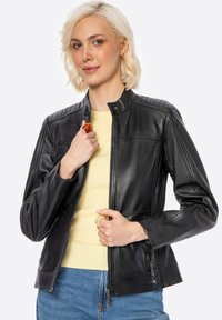 Giubbotto in pelle nera con accenti trapuntati sulle spalle, colletto alto, zip frontale e tasche laterali. Indossato sopra una maglia gialla, abbinato a jeans blu.