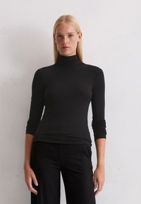 Blonde Frau, die einen schwarzen Langarmrollkragenpullover und schwarze Hosen trägt, steht vor einer schlichten weißen Wand und blickt nach vorne.