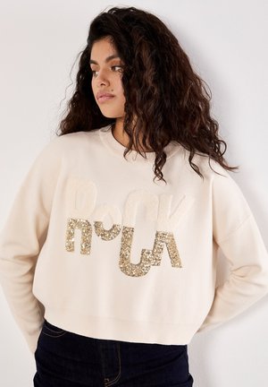 Sweatshirt court couleur crème avec des lettres « Rock » en sequins dorés et tissu peluche. Il possède un col rond et des poignets côtelés.