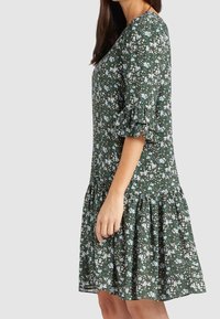 Vestido floral verde con forma acampanada, que presenta mangas con volantes, patrones de flores en blanco y azul, y un dobladillo en capas. Tela ligera.