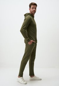 Olivengrünes Hoodie- und Jogginganzug-Set aus weichem Stoff. Kapuzen-Design mit seitlichen Taschen; kombiniert mit weißen Sneakern für einen lässigen Look.