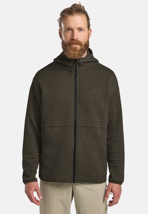 Mann mit rotem Bart und zurückgebundenen Haaren, der einen dunkelolivgrünen, gesteppte Kapuzenjacke mit Reißverschluss und hellbeige Hose trägt, vor einfarbigem Hintergrund steht.