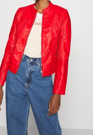 Personne portant une veste zippée rouge vif, un t-shirt rayé blanc et jaune, et un jean bleu taille haute sur un fond uni.