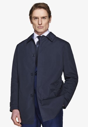 Mężczyzna w granatowym płaszczu na niebieskim garniturze, białej koszuli i ciemnym krawacie, stojący z jedną ręką w kieszeni na jednolitym tle.