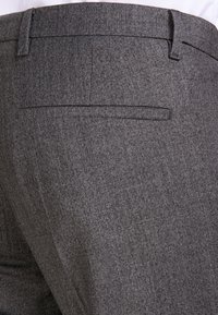 Primo piano di pantaloni grigi texturizzati con una tasca a filetto sul retro e passanti per cintura, indossati con una camicia bianca.
