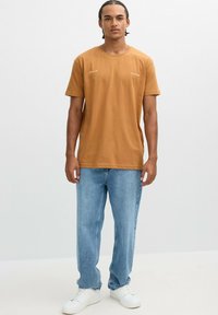 Camiseta de algodón marrón con mangas cortas y escote redondo, combinada con jeans de mezclilla azul claro y zapatillas blancas. Diseño simple, sin patrones.