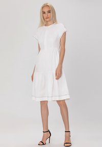 Robe en coton blanche à manches courtes, jupe plissée, encolure ronde et couture décorative le long de l'ourlet ; assortie avec des talons noirs.