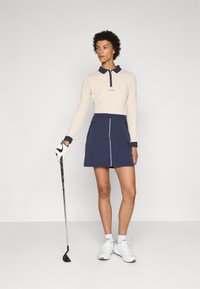 Long-sleeve beige polo with navy collar, paired with a navy skirt featuring white trim. Model holds a golf club and wears white sneakers.
