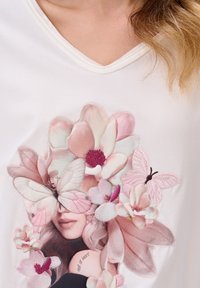 Witte V-hals T-shirt met een geborduurd ontwerp van roze bloemen en vlinders, met een gedeeltelijk zichtbare gezicht en de tekst "echt van binnen."
