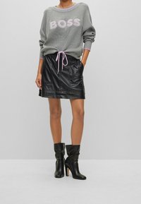 Grauer Strickpullover mit pinkem "BOSS"-Logo, kombiniert mit einem schwarzen Lederrock mit elastischem Bund und zwei Vorderaschen. Schwarze Knöchelstiefel.