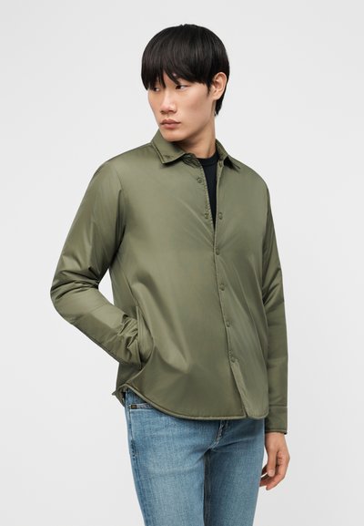 Jeune homme portant une veste boutonnée vert olive et un jean bleu, debout avec une main dans la poche, regardant sur le côté.