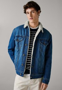 Jeansjacka med en blå utsida, vit fuskpälskrage, tryckknappar och två bröstfickor. Bärs över en mörkrandig tröja.