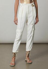 Pantalones cargo blancos de talle alto, con pliegues, cinturón de lazo y dos bolsillos laterales. Combinados con sandalias beige de tiras con acentos dorados.