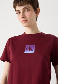 Burgunderfarget bomulls t-skjorte med korte ermer, med et kvadratisk grafikk i lilla og rødt med teksten "cK" sentrert.