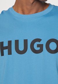 Jasnoniebieska bawełniana koszulka z odważnym czarnym logo "HUGO" z przodu. Okrągły dekolt i miękka tekstura. Design jest minimalistyczny i nowoczesny.