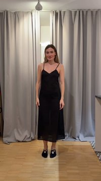 Γυναίκα που στέκεται σε εσωτερικό χώρο σε ξύλινο πάτωμα, φοράει μαύρο μακρύ φόρεμα χωρίς μανίκια και μαύρες μπαλαρίνες, με κλειστές μπεζ κουρτίνες πίσω της.