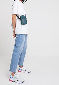 Tyrkysová crossbody taška s černým popruhem, nošená přes bílé tričko. Světle modré džíny a bílé tenisky s růžovými a modrými akcenty.