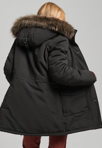 Black parka coat with a fur-lined hood, featuring a loose fit, quilted texture, and front pocket. Soft fur trim and adjustable hood detail.