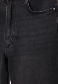 Gros plan sur un jean en denim noir montrant la poche avant, la couture et un rivet en métal sur un tissu texturé.