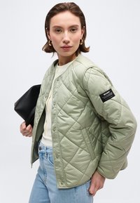 Γυναίκα με ανοιχτό πράσινο μπουφάν καπιτονέ με μαύρη ετικέτα ECOALF στο μανίκι, λευκό τοπ, ανοιχτό μπλε τζιν, κρατώντας μαύρη τσάντα φάκελο.