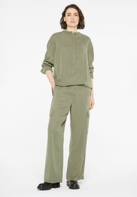 Olivgrünes Outfit mit einer locker sitzenden, knopfbaren Bluse mit Stehkragen und weiten Cargo-Hosen. Der Stoff hat eine weiche Textur. Schwarze Stiefel.