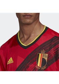 Maglia rossa con dettagli neri, scollatura a V con rifiniture gialle. Presenta il logo della nazionale belga e il marchio Adidas sulla spalla.