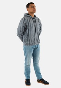 Chaqueta con capucha en gris claro con un patrón gráfico negro, combinada con jeans de mezclilla azul claro y zapatillas oscuras con acentos marrones.